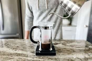 Home barista pouring water from a gooseneck kettle into a French press sitting on a kitchen scale